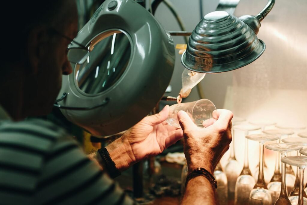 An artisan carefully shaping a glass piece under magnifying lamp indoors.