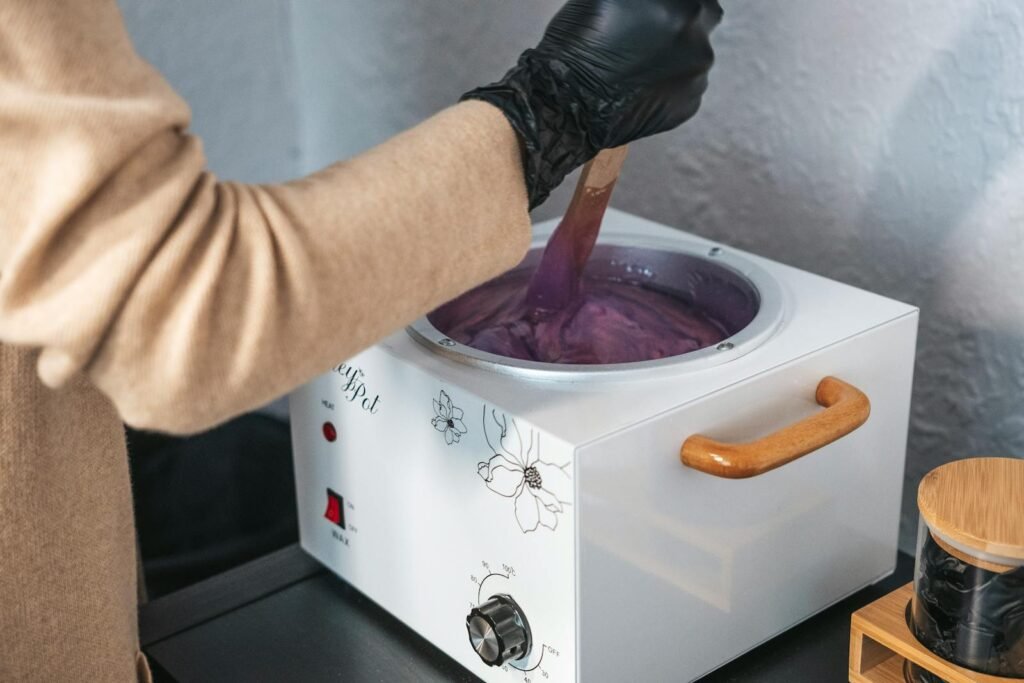 An esthetician wearing gloves stirring purple hair wax in a wax pot indoors